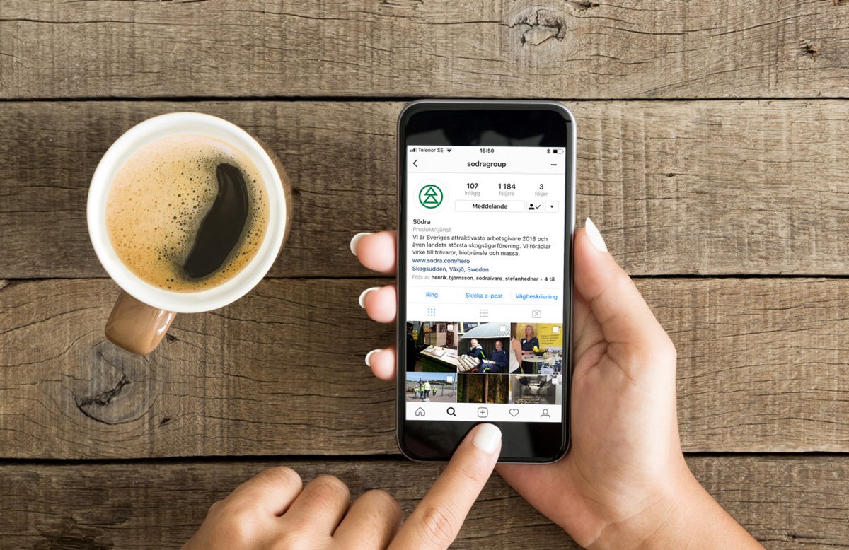 Kaffemugg med kaffe. Två händer. Höger hand håller i smartphone med instagram. Vänsterhandens pekfinger trycker på hemknappen. Gammalt träbord i bakgrunden. Bilden tagen ovanifrån.