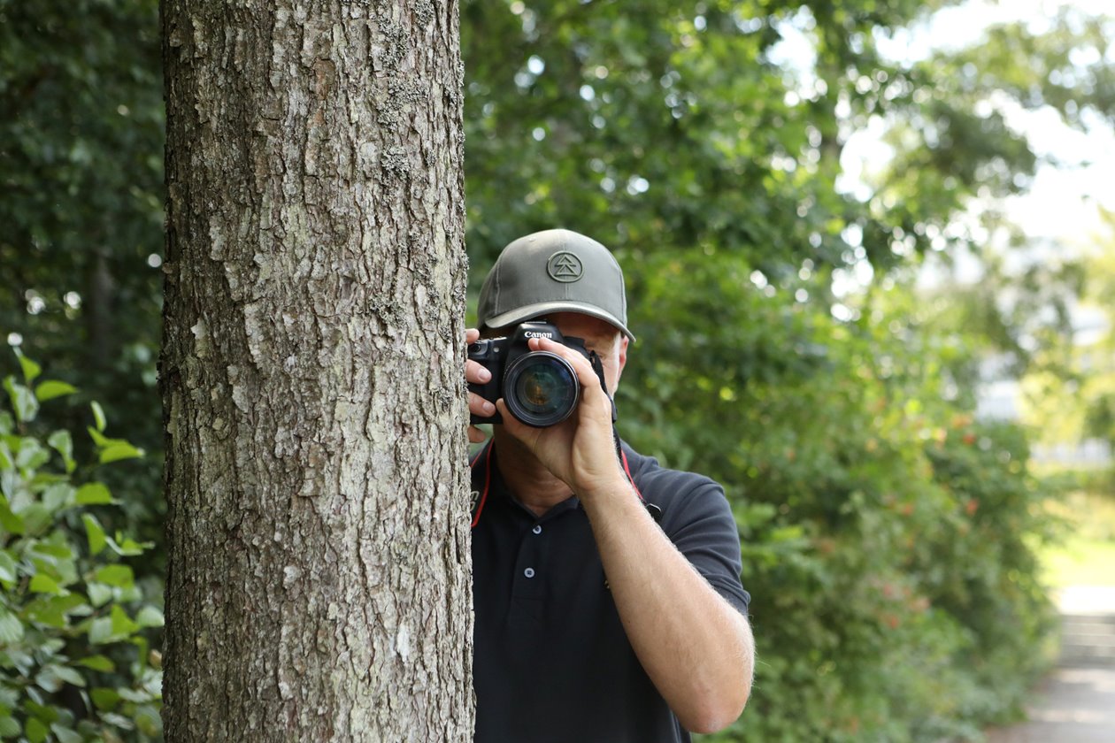 Fotograf står bakom träd och håller i kamera.