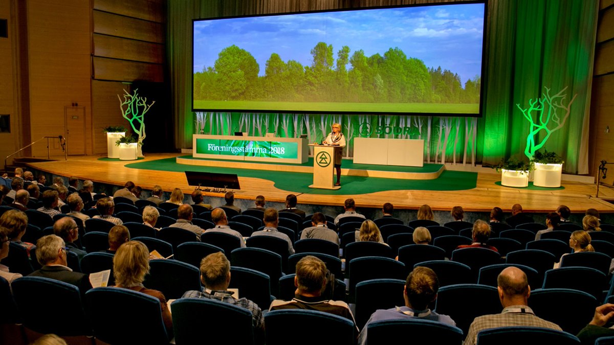 Stor scen med trägolv, grön matta, presidiebord och talarstol i trä. Grön banderoll med vit text "Föreningsstämma 2018". Stor bildskärm med lövskog. Södras ordförande i talarstolen. I förgrunden många människor som sitter i långa rader på blå stolar med ryggen mot kameran.