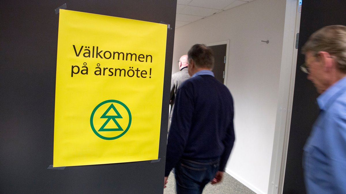 Gul skylt med grön Södrasymbol. Text: Välkommen på årsmöte! Tre män går in i mötelokalen till höger om skylten.