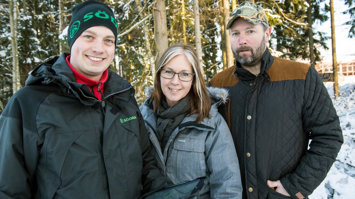 Tre personer står ute i vinterskog. Till vänster ung skogsinspektor med svart Södrajacka, svart Södramössa och surfplatta i handen. I mitten kvinna i grå jacka, glasögon, brunt långt här. Till höger man med brun jacka, kamouflagekeps, skägg.