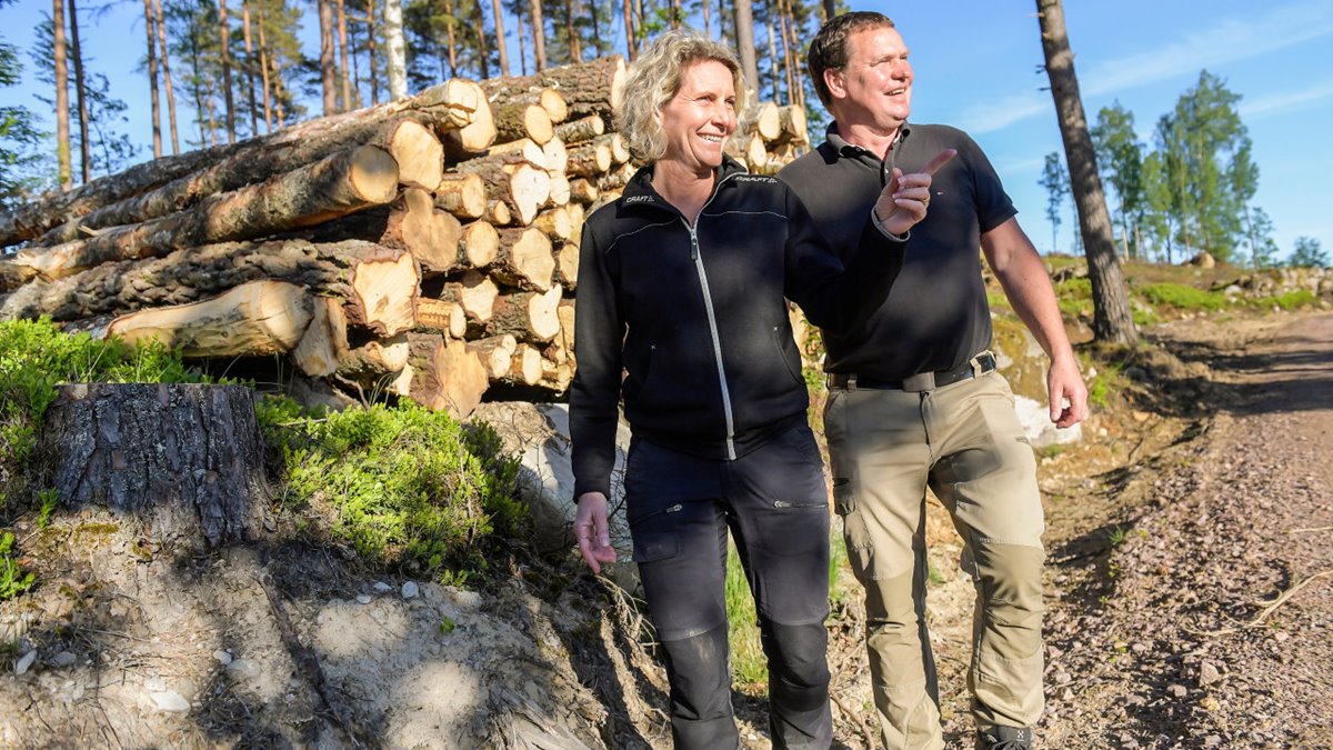 Medelålders par på skogsväg framför virkestrave. Kvinnan har ljust lockigt halvlångt här, svart jacka och svarta byxor. Hon ler och pekar på något till höger utanför bilden. Mannen till höger om henne har mörtt hår, svart tenniströja och beige byxor. Han ler och tittar åt det håll som kvinnan pekar. Soligt. Blå himmel. Trädstammar i bakgrunden.