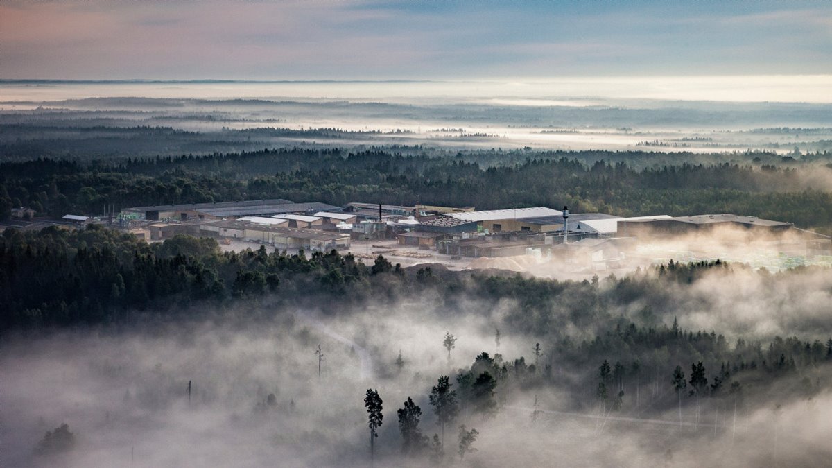 Sågverket i Långasjö från luften, fotot taget snett uppifrån. Soligt. Dimma över vissa markpartier. Sågverksbyggnaden i mitten. Skog. Blå himmel.