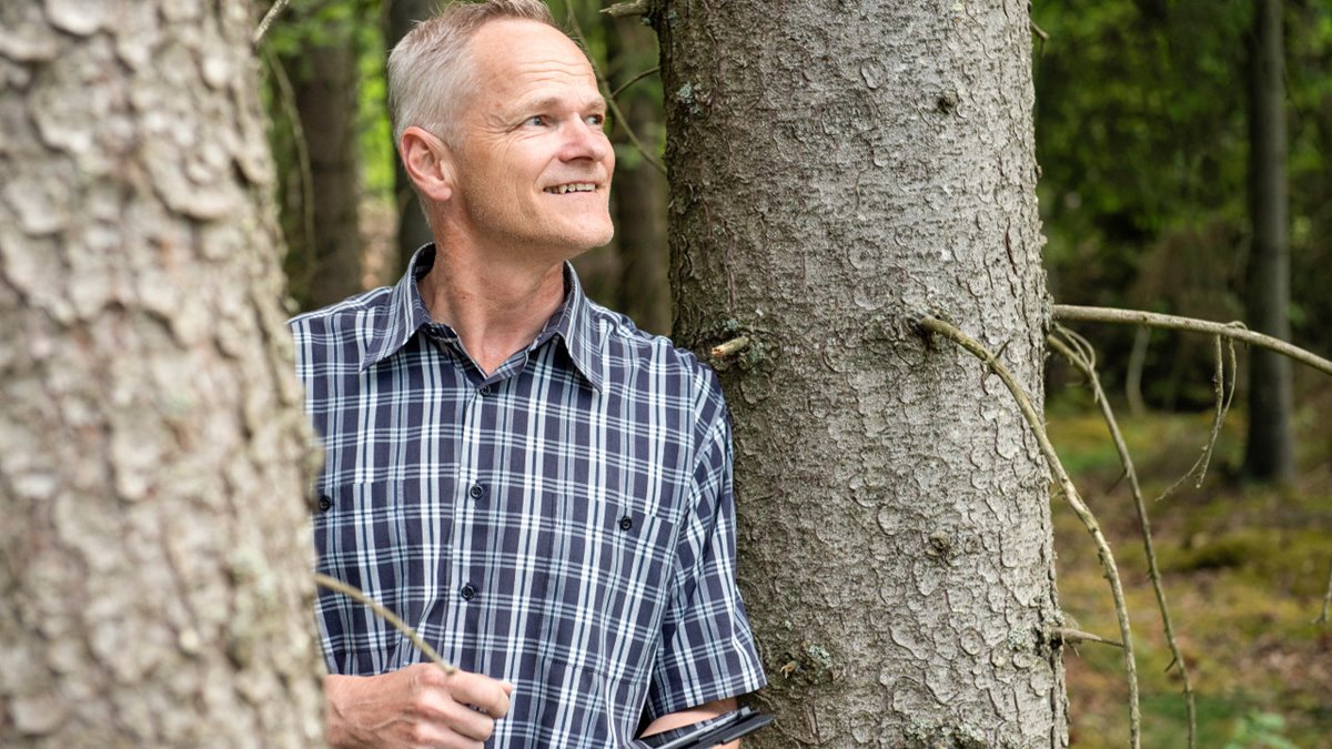Man står mellan två granstammar. Grått hår, blå- och vitrutig, kortärmad skjorta. Håller en surfplatta i ena handen. Tittar snett uppåt höger. Ler.