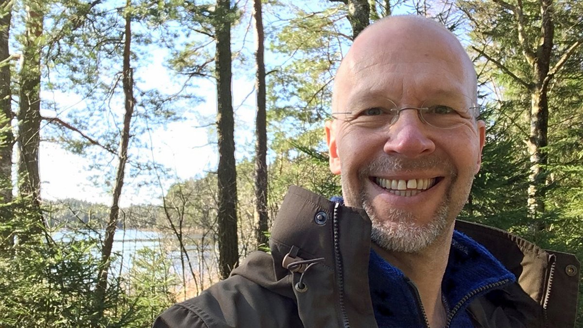 Medelålders man i brun jacka med blå tröja under. Glad, glasögon, grått skägg, inget hår på huvudet. Lövskog och sjö i bakgrunden. Soligt.