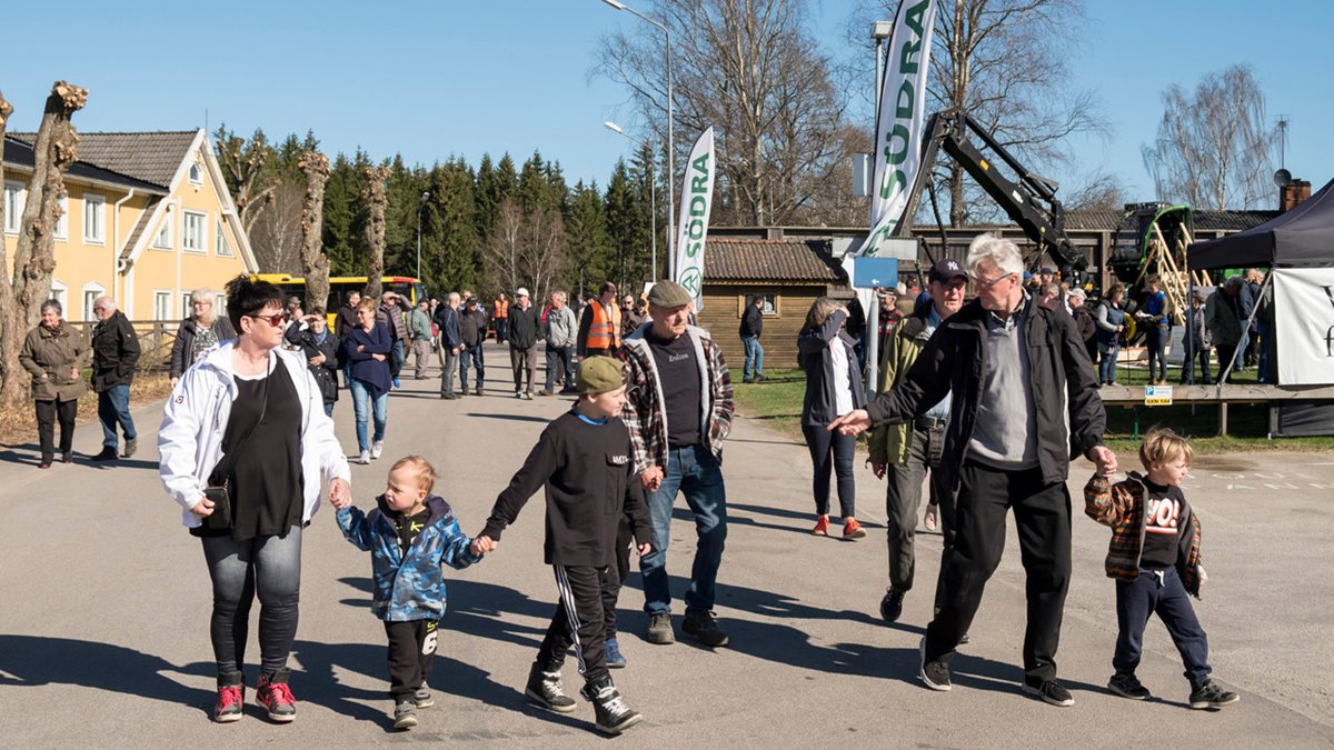 Många människor går in på sågverksområde. I bakgrund gult hus, granar, avverkningsmaskin, beachflaggor med Södra-loggor. I förgrunden till vänster en kvinna med mörkt kort hår och vit jacka. Håller två barn i handen. I mitten en medelålders man med keps och rutig jacka. Till höger en man med grått hår, glasögorn, svart jacka, grå tröja och svarta byxor. Håller lite pojke i handen.