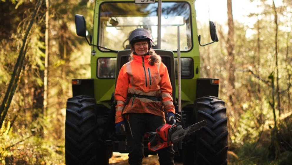 Kvinna framför traktor i skyddsutrustning och motorsåg