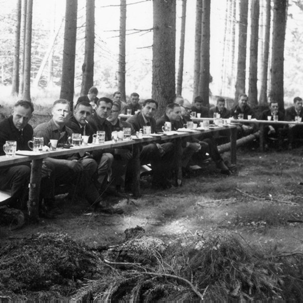 Svart vit bild av samling av människor som sitter i skogen vid långbord