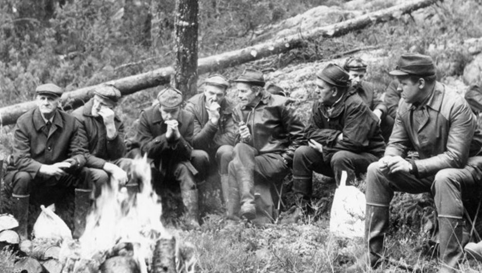 Män som sitter i skog vid en eld.